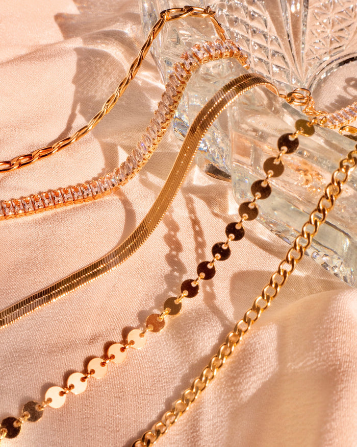 Five different gold bracelets laying draped across a glass dish on a pink background