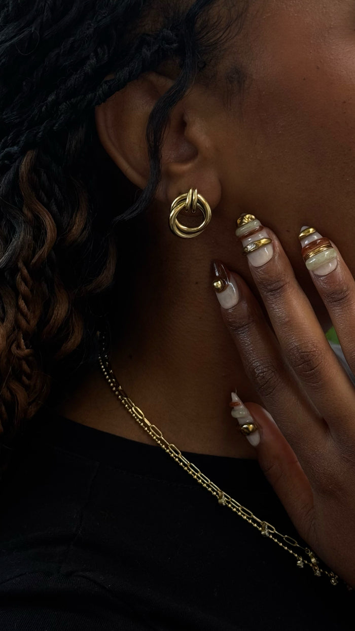 Side shot of a woman with curly hair wearing statement earring, with accented metallic nails and gold necklaces