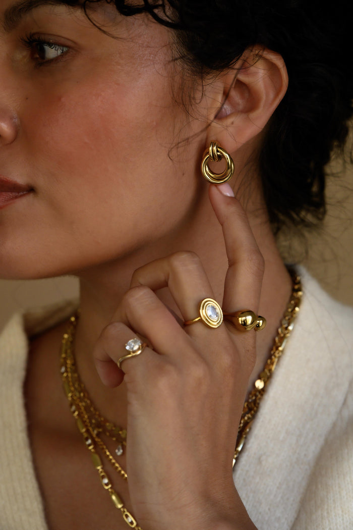 Close-up side view of a woman wearing gold statement earrings, rings, and necklaces in a white sweater