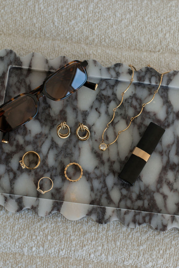 Flatlay of sunglasses, rings, earrings, a necklace, and lipstick on a grey marble tray set on a white fabric background