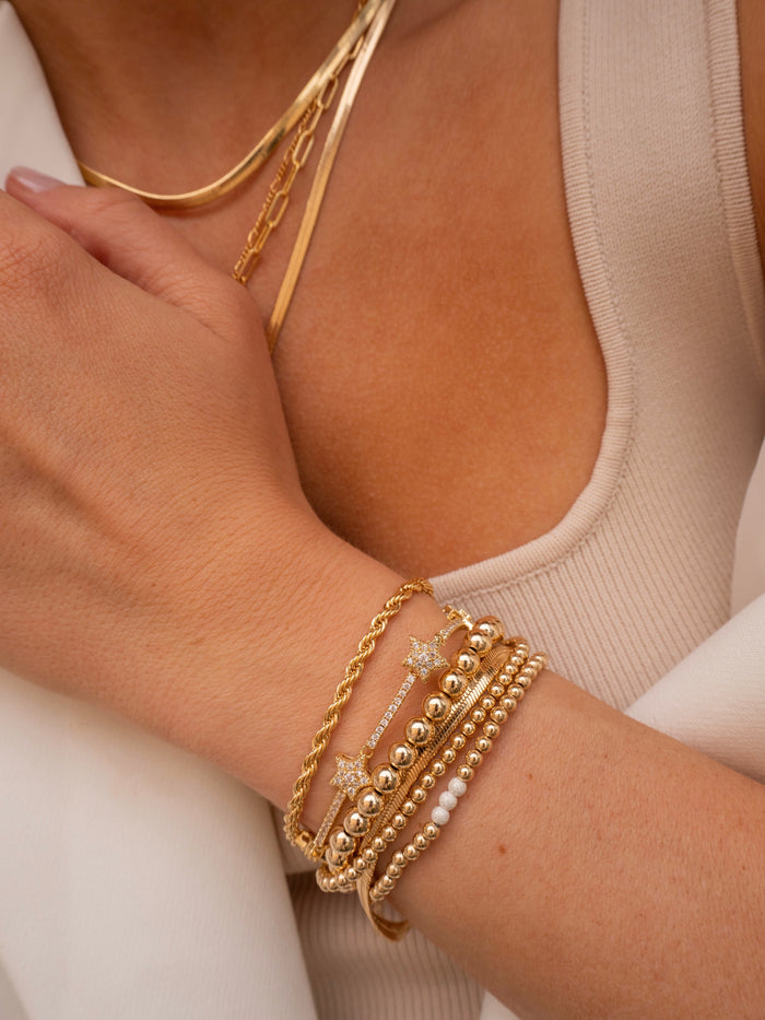 A person wearing a tan tank top, white pants, gold necklaces, and gold and stone bracelets