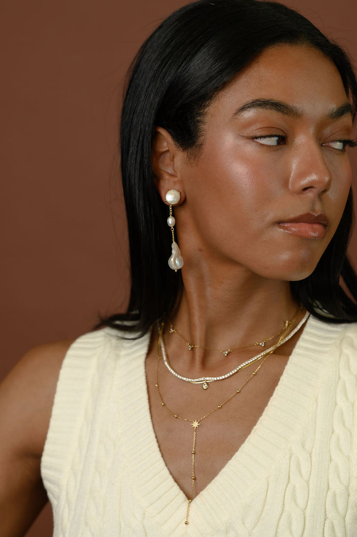 A woman wearing gold and pearl jewelry against a brown background