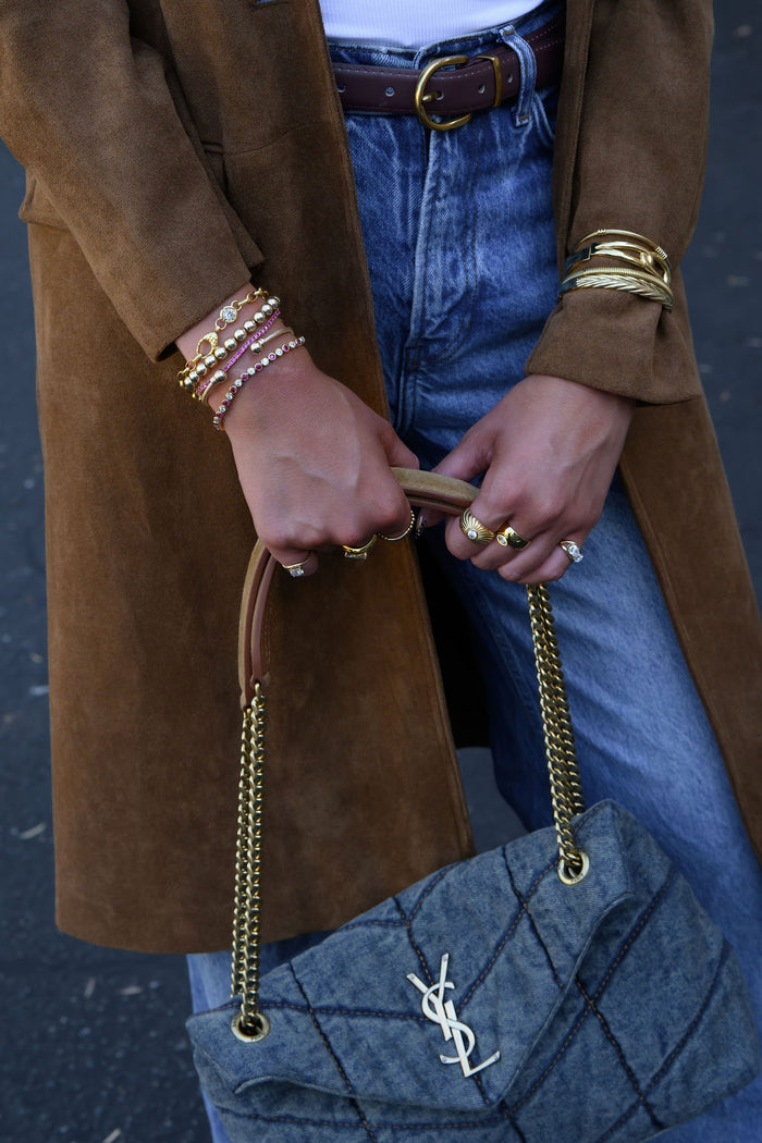 A person wearing a brown coat, blue jeans, and holding a purse, with gold rings and bracelets