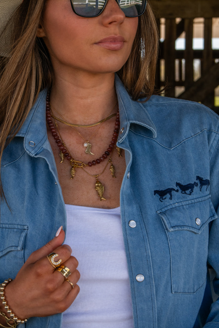 Woman wearing a blue denim jacket with horse designs, sunglasses, and layered necklaces.
