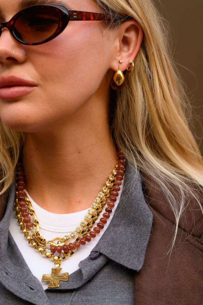 A woman wearing gold jewelry and sunglasses against a brown wall