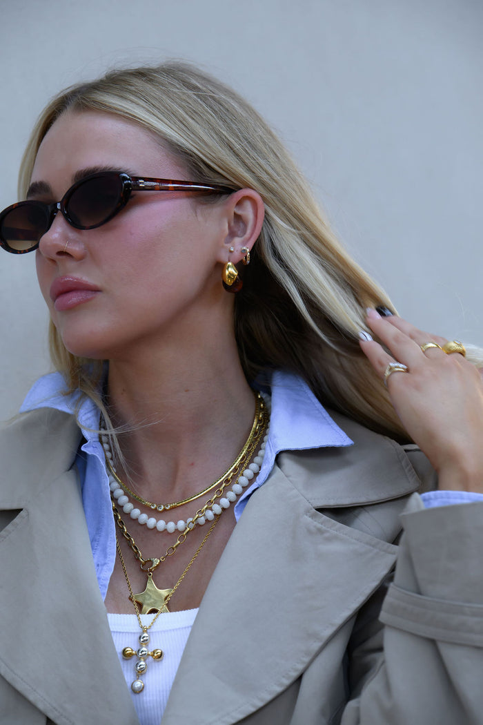 A woman wearing gold jewelry and sunglasses in a tan trench coat against a white wall