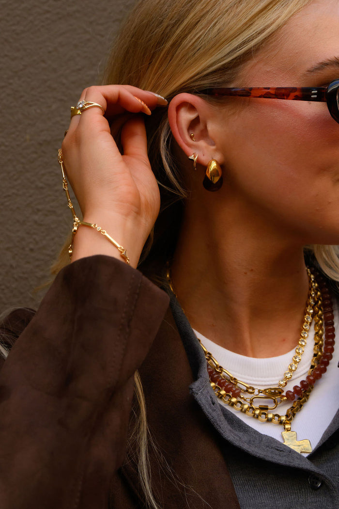 A woman wearing gold jewelry including earrings and a hand chain and sunglasses against a brown wall