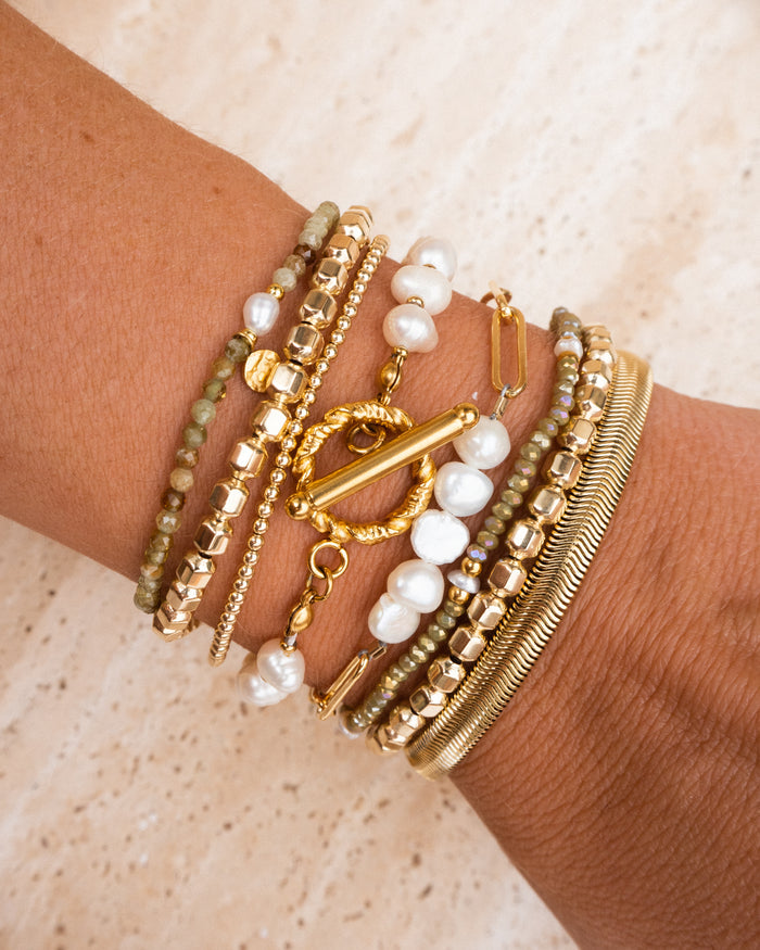 A wrist with gold and pearl bracelets on a tan stone background