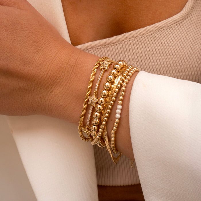 Close up of a person's chest and wrist wearing a white blazer, tan top, and assorted gold bracelets