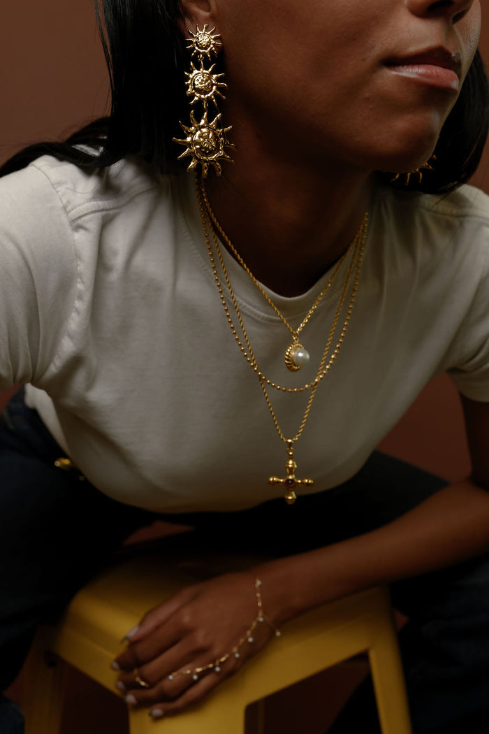Woman wearing statement gold earrings and necklaces leaning on a yellow stool
