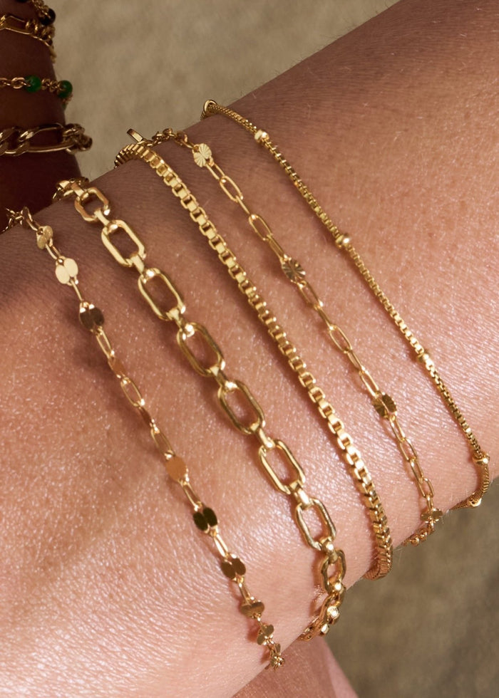Close up of two wrists with dainty gold chain bracelets on a tan background