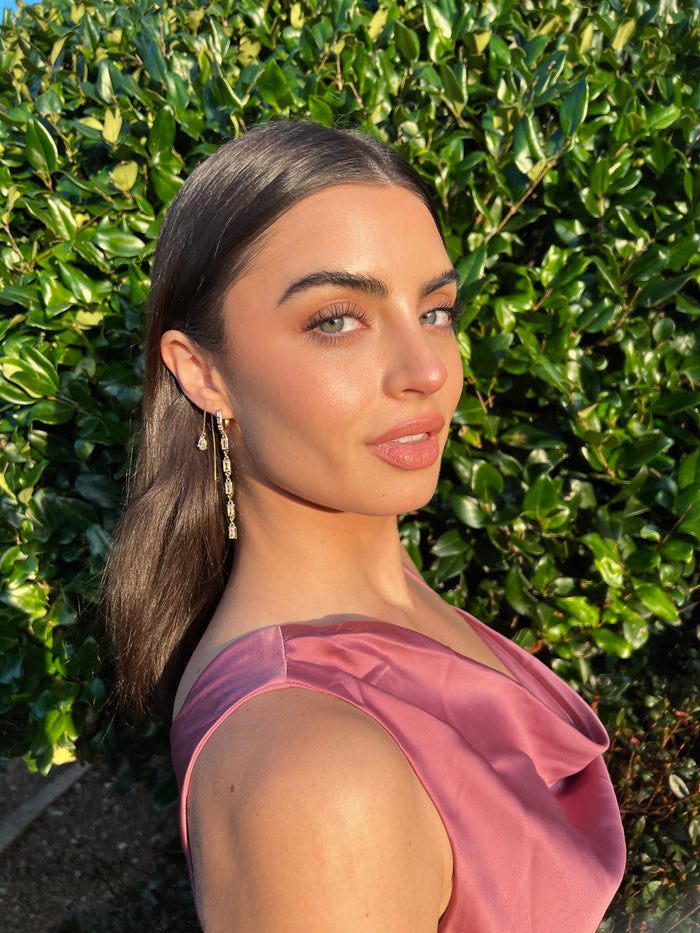 A woman in a pink dress and stone earrings posing in front of greenery