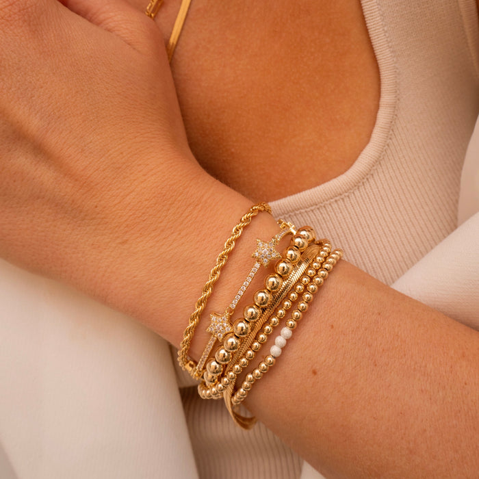 A person wearing a tan tank top, white pants, gold necklaces, and gold and stone bracelets