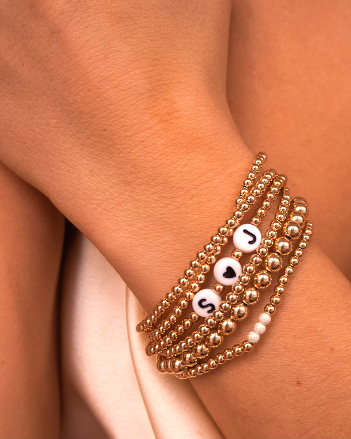 Close up of a wrist with gold bead bracelets, including a custom bead bracelet that reads "S<3J", on a white background