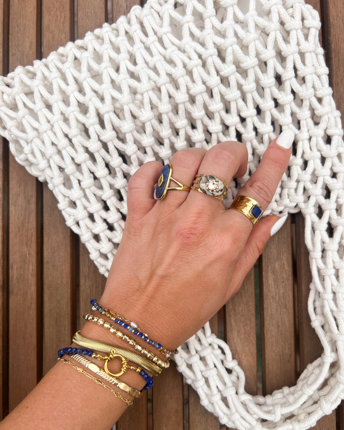 A hand with gold and blue rings and bracelets on a wooden bench with a white crochet purse
