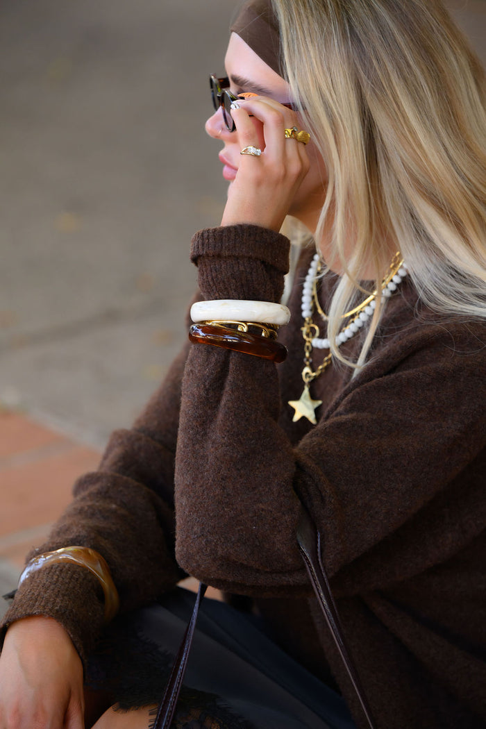 A woman wearing a brown sweater, sunglasses, and various gold and stone jewelry