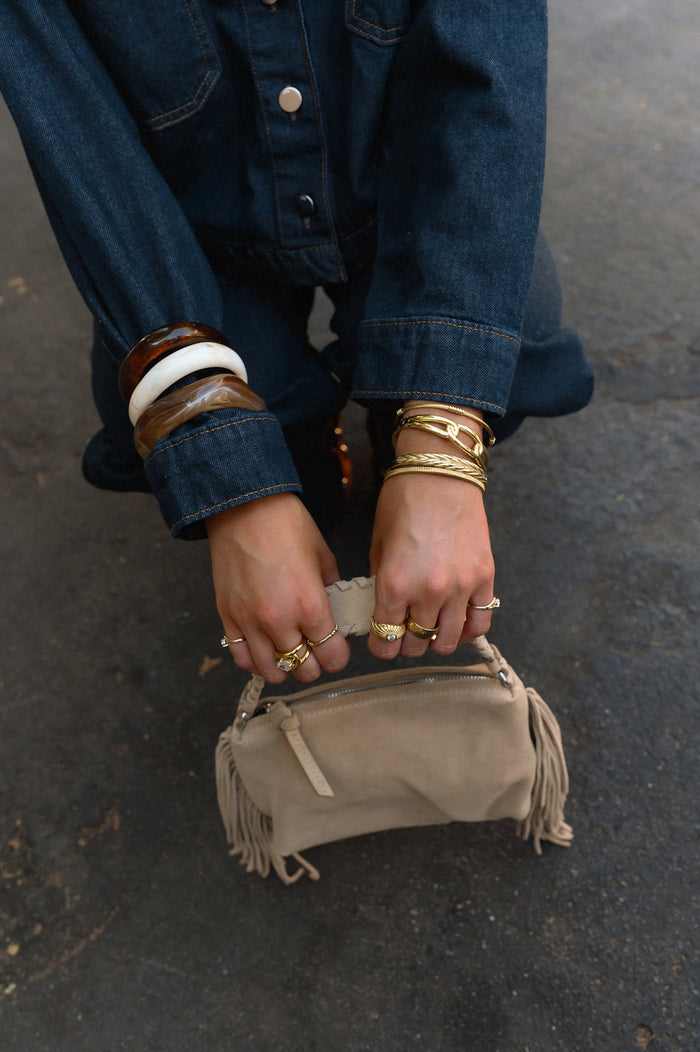 A person in a denim set wearing gold and stone bracelets and rings holding a purse